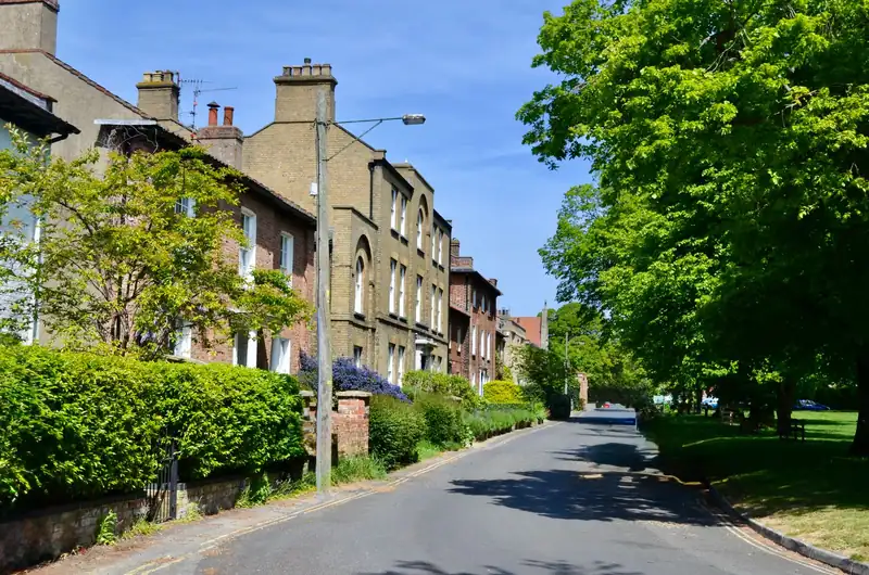 Brick terrace with low front walls and hedges along a kerbed pavement, the kind of footway a vehicle crossing has to be cut through in Royal Wootton Bassett